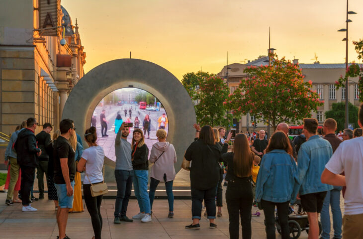 A group of people gather in front of a Portal artwork to get a glimpse of onlookers in a different location.