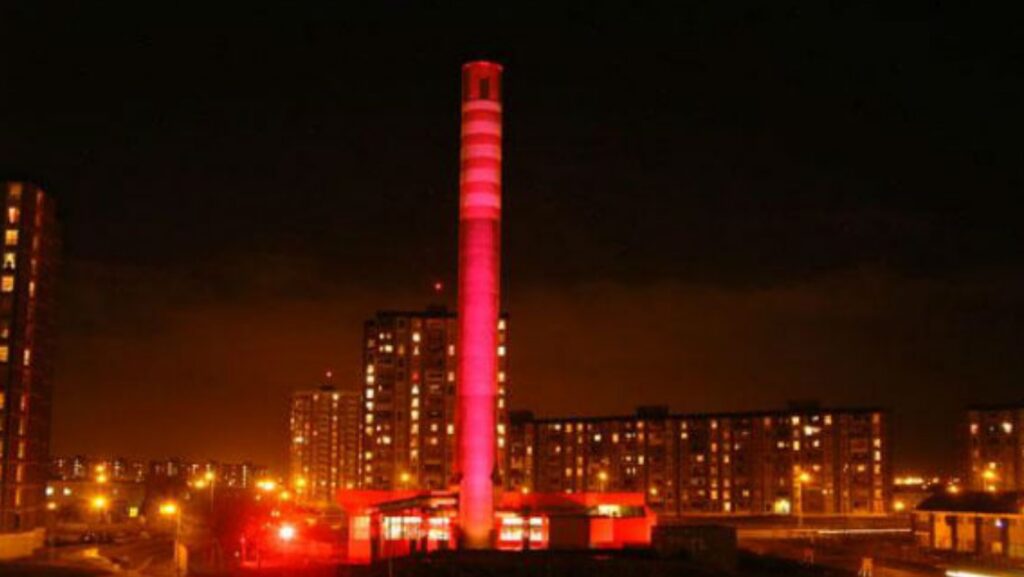 A chimney floodlit in pink light with city lights at night in background