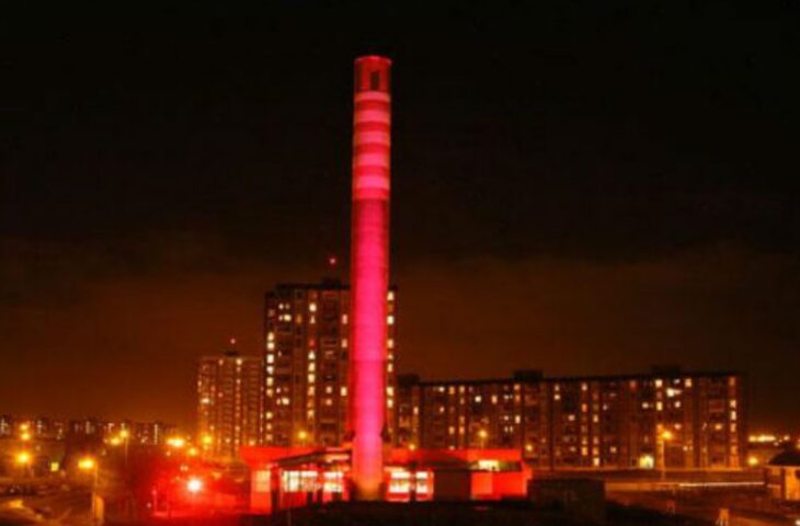 A chimney floodlit in pink light with city lights at night in background
